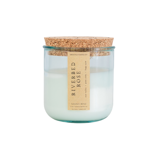 A glass jar candle with a cork lid and a beige paper label reading 'RIVERBED ROSE' on a plain white background.