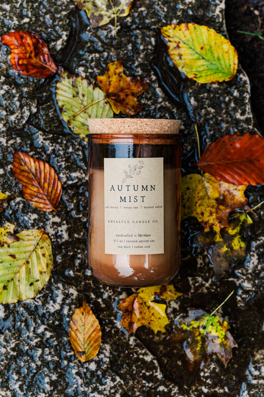 Autumn Mist candle by Unsalted Candle Co. surrounded by rain-soaked fall leaves on wet rock.