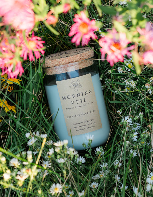 Morning Veil floral candle by Unsalted Candle Co. nestled in green meadow grass surrounded by pink wildflowers and delicate white daisies.