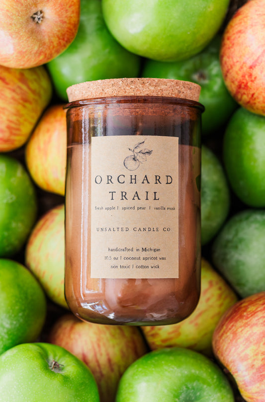 Orchard Trail apple scented candle by Unsalted Candle Co. in an amber glass jar surrounded by fresh green and red apples.