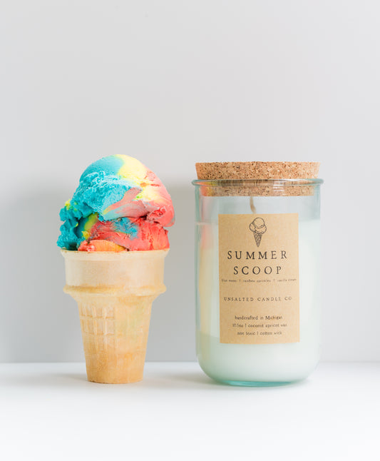 A jar candle labeled 'SUMMER SCOOP' by Crafted Candle Co stands next to a colorful ice cream scoop in a waffle cone against a clean light gray background.
