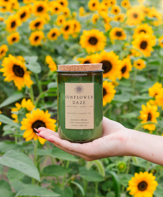 A hand holds a green glass jar candle with a cork lid and a cream label reading 'Sunflower Daze' in front of a blurred field of yellow sunflowers.