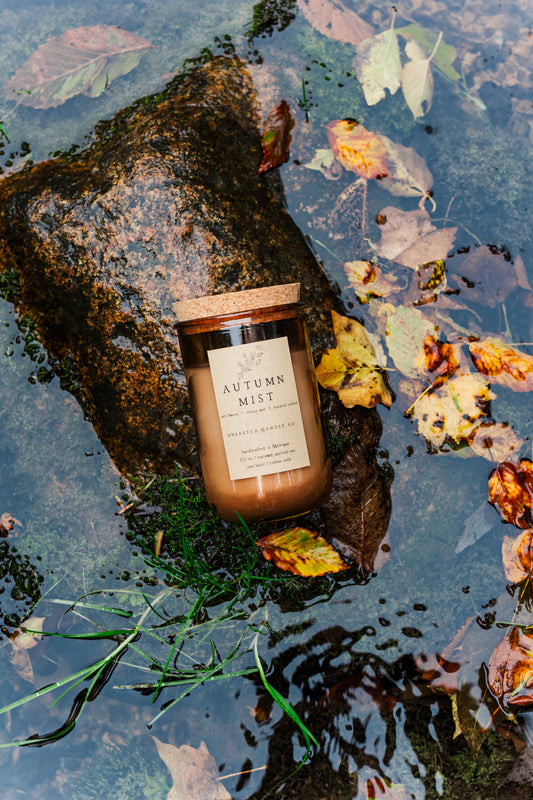 Autumn Mist fall candle by Unsalted Candle Co. beside wet autumn leaves and moss-covered rocks in rainwater.