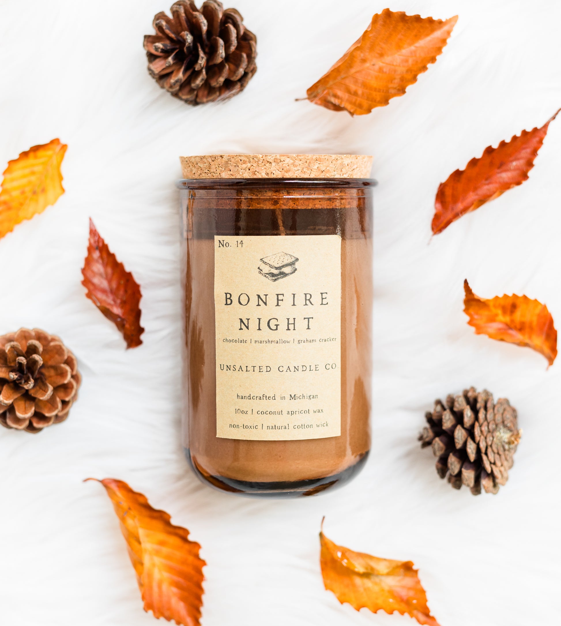 A glass jar candle with a cork lid and a beige label reading 'BONFIRE NIGHT' and 'UNSALTED CANDLE CO' is arranged on a white textured surface with autumn leaves and pinecones around it.