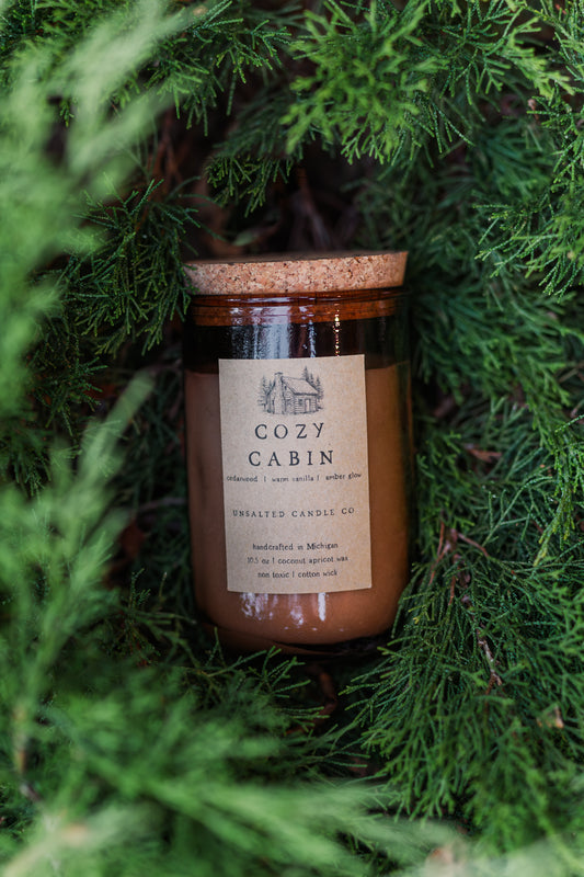 Cozy Cabin candle by Unsalted Candle Co. nestled among lush evergreen branches, featuring cedarwood, warm vanilla and amber notes.