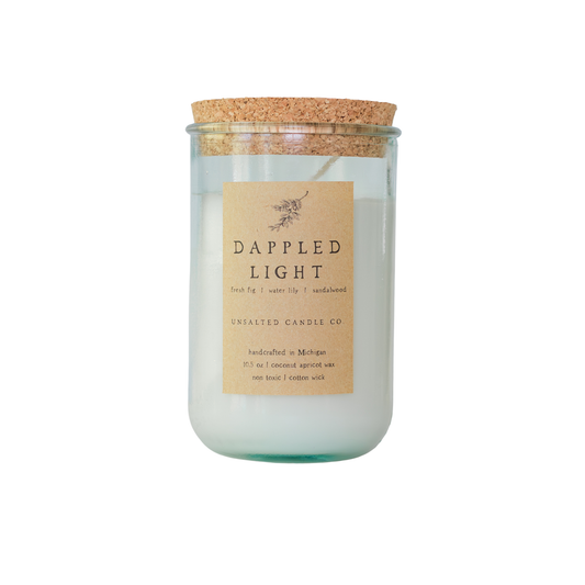 Clear glass jar candle filled with white wax and capped with a natural cork lid. The tan paper label reads 'DAPPLED LIGHT' and 'UNSALTED CANDLE CO.' on a plain white background.