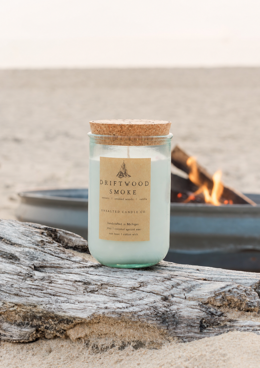 Driftwood Smoke candle by Unsalted Candle Co. resting on weathered driftwood with a glowing beach bonfire in the background.