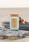 Driftwood Smoke candle by Unsalted Candle Co. resting on weathered driftwood with a glowing beach bonfire in the background.