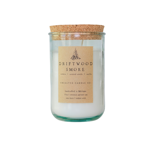 Clear glass jar candle with white wax and a natural cork lid. The tan paper label features a campfire illustration and reads 'DRIFTWOOD SMOKE', 'embers | smoked woods | vanilla', and 'UNSALTED CANDLE CO.' on a white background.