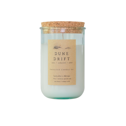 Clear glass jar candle with white wax and a natural cork lid. The tan paper label features a sand dune illustration and reads 'DUNE DRIFT', 'rattan | sandalwood | amber', and 'UNSALTED CANDLE CO.' on a white background.