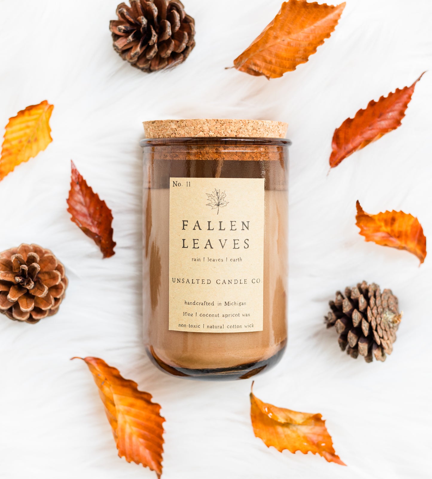 A jar candle labeled 'Fallen Leaves' by Unsalted Candle Co, styled in a flat lay with autumn leaves and pinecones on a white textured background.