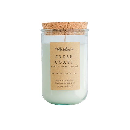 Clear glass jar candle with white wax and a natural cork lid. The tan paper label reads 'FRESH COAST', 'mineral air | wet stone | driftwood', and 'UNSALTED CANDLE CO.' on a white background.