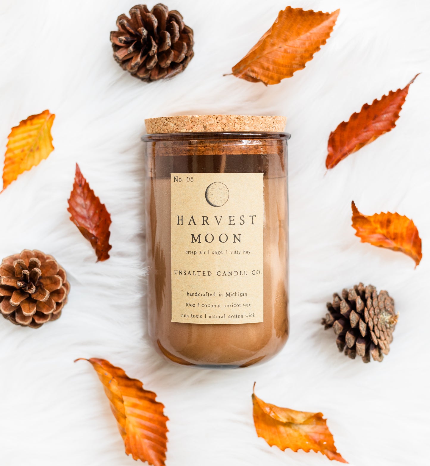 A jar candle labeled 'Harvest Moon' by Unstalted Candle Co, styled in a flat lay with autumn leaves and pinecones on a white textured background.