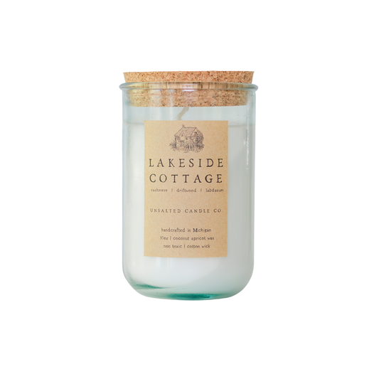 Clear glass jar candle with white wax and a natural cork lid. The tan paper label features a cottage illustration and reads 'LAKESIDE COTTAGE', 'cashmere | driftwood | labdanum', and 'UNSALTED CANDLE CO.' on a white background.