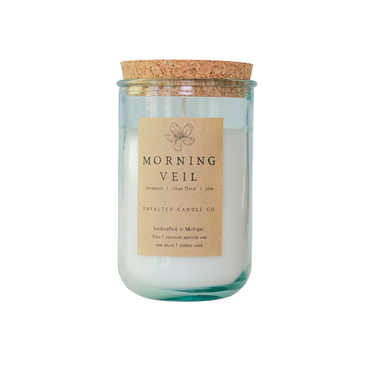 Clear glass jar candle with white wax and a natural cork lid. The tan paper label features a flower illustration and reads 'MORNING VEIL', 'bergamot | clean floral | pear', and 'UNSALTED CANDLE CO.' on a white background.