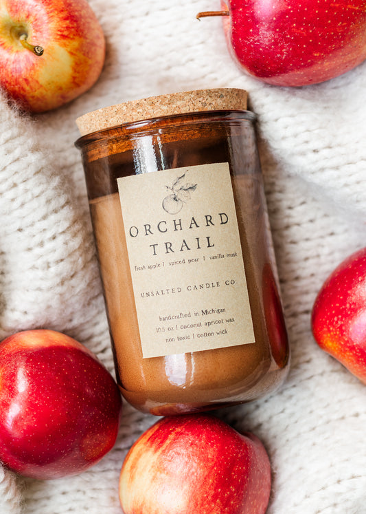 Orchard Trail candle by Unsalted Candle Co. surrounded by fresh red apples on a cozy knit blanket.