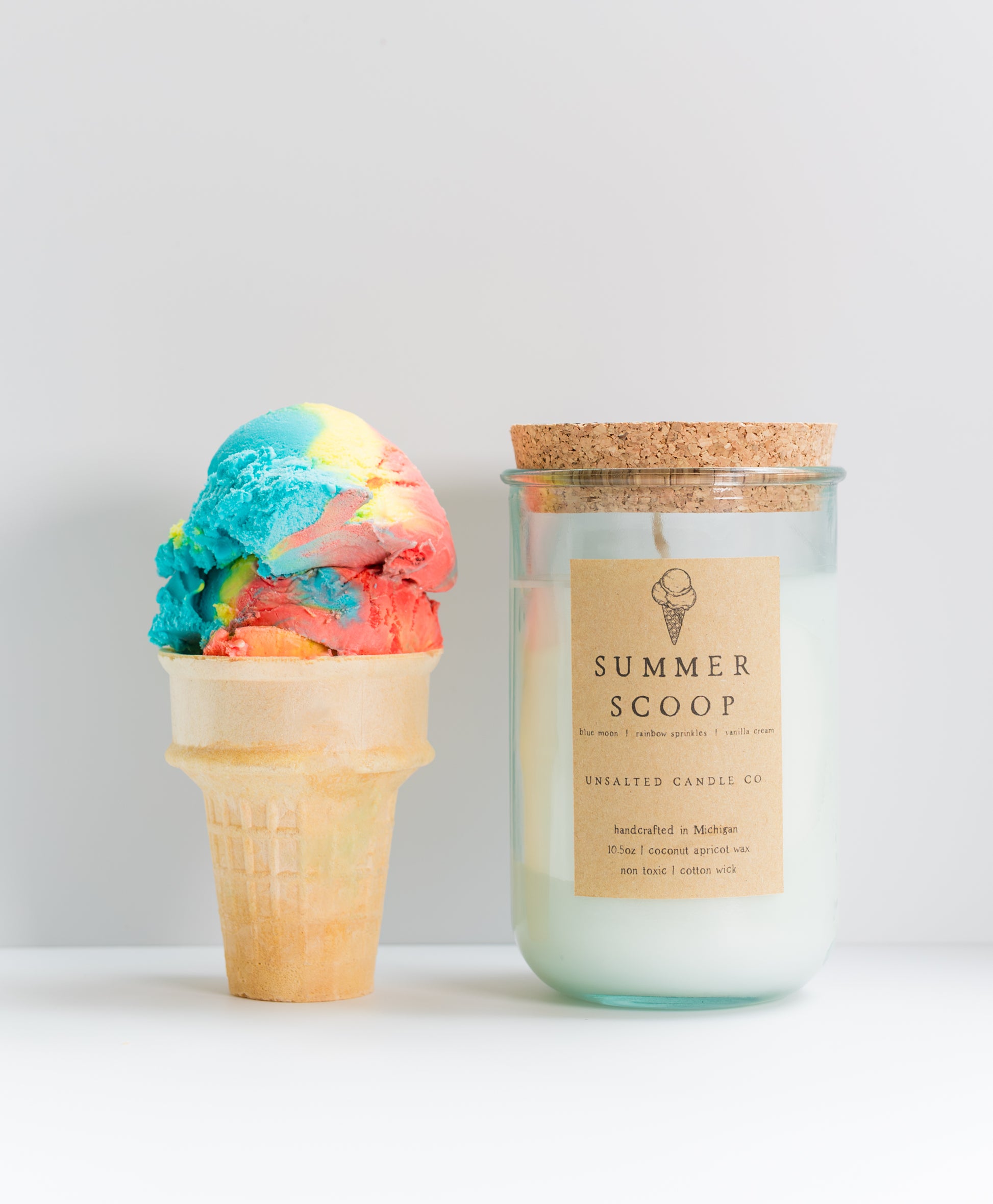 A jar candle labeled 'SUMMER SCOOP' by Crafted Candle Co stands next to a colorful ice cream scoop in a waffle cone against a clean light gray background.