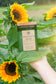 A hand holds a green glass jar candle labeled 'Sunflower Daze' in front of sunflowers and green leaves outdoors.