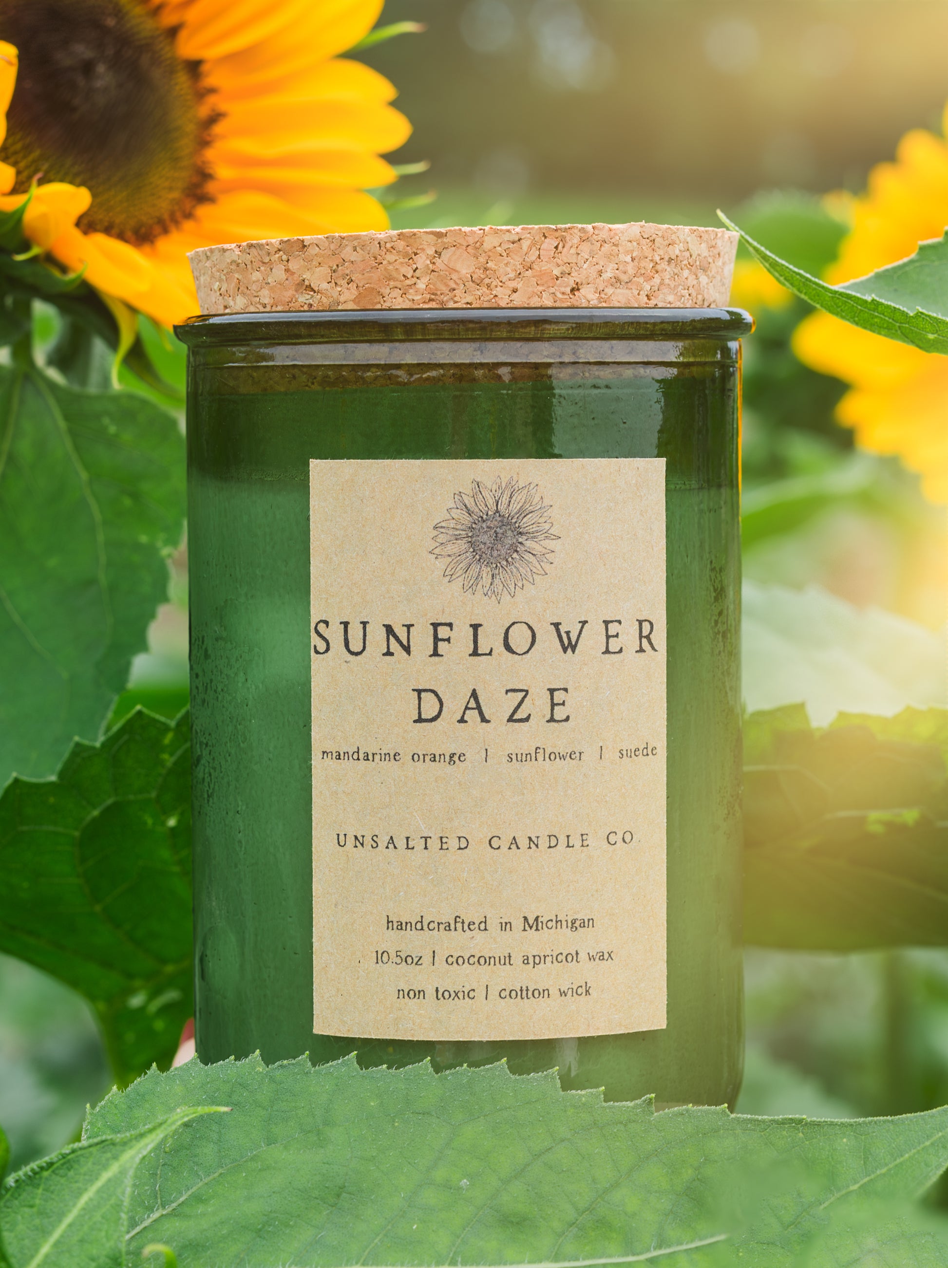 A jar candle labeled 'Sunflower Daze' by Unsalted Candle Co, set among green leaves and bright yellow sunflowers. The candle has a dark green glass jar with a natural cork lid and a beige paper label.