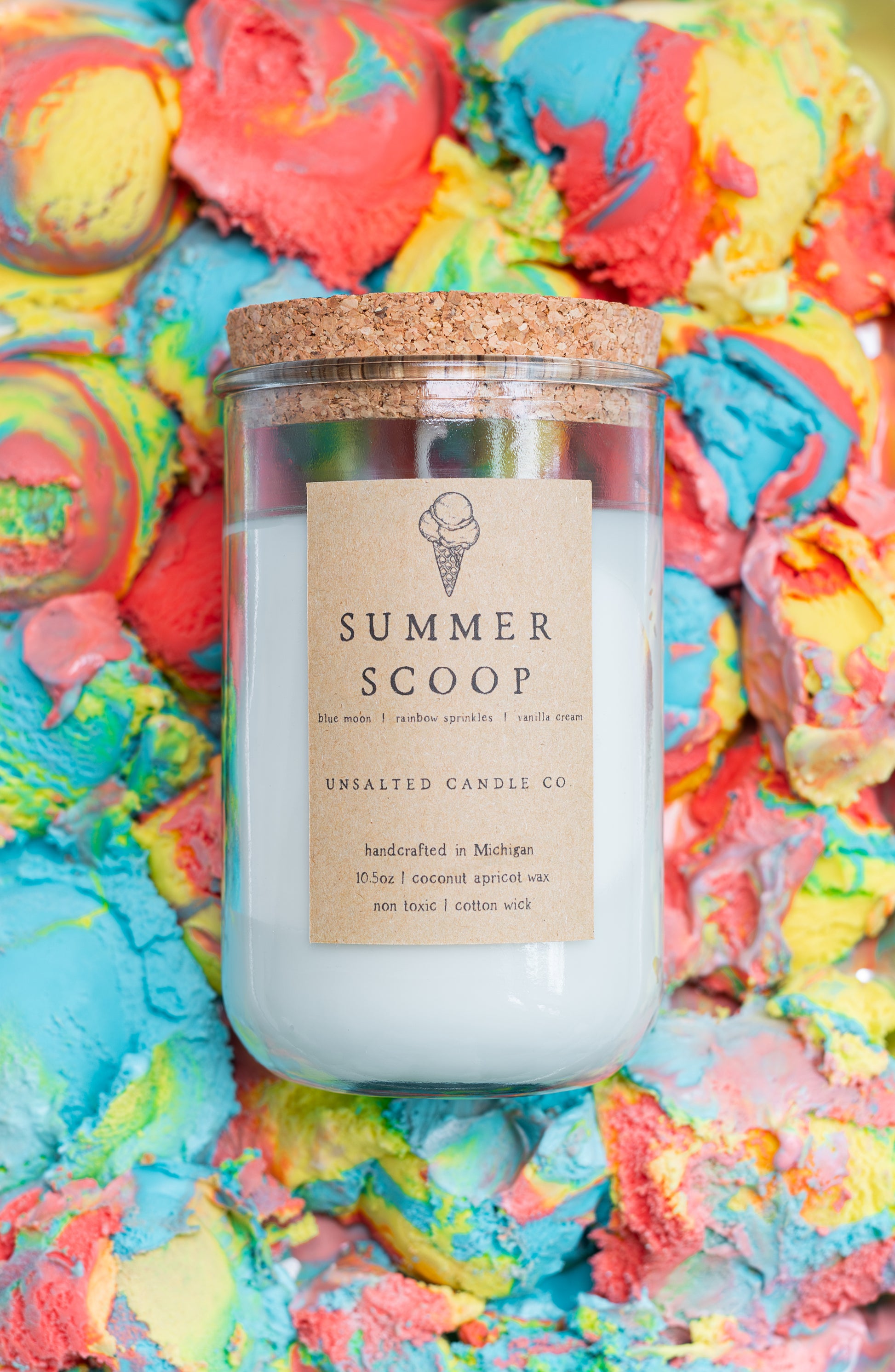 A glass jar candle with a cork lid and a kraft paper label reading 'SUMMER SCOOP' and 'UNSALTED CANDLE CO.' The candle is placed on top of a colorful, textured background resembling melted ice cream swirls in blue, pink, yellow, and orange.
