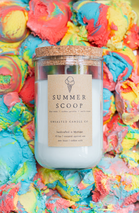 A glass jar candle with a cork lid and a kraft paper label reading 'SUMMER SCOOP' and 'UNSALTED CANDLE CO.' The candle is placed on top of a colorful, textured background resembling melted ice cream swirls in blue, pink, yellow, and orange.