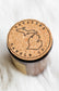 A close-up top-down view of a candle jar with a round cork lid printed with the brand name 'UNSALTED CANDLE CO.' and a map outline of Michigan, resting on a white faux fur surface.